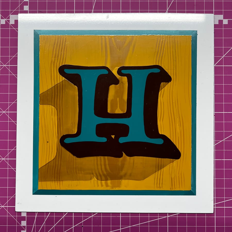 A teal letter H with a hard cast shade, painted on a grained ochre panel