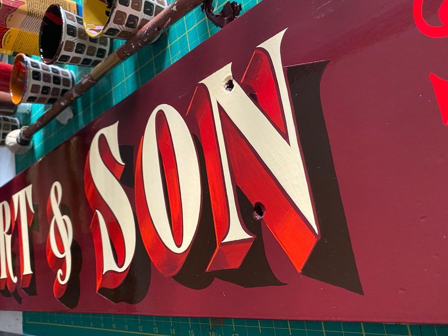 A red sign panel hand-lettered in cream with a deep cast shade, on the workshop bench